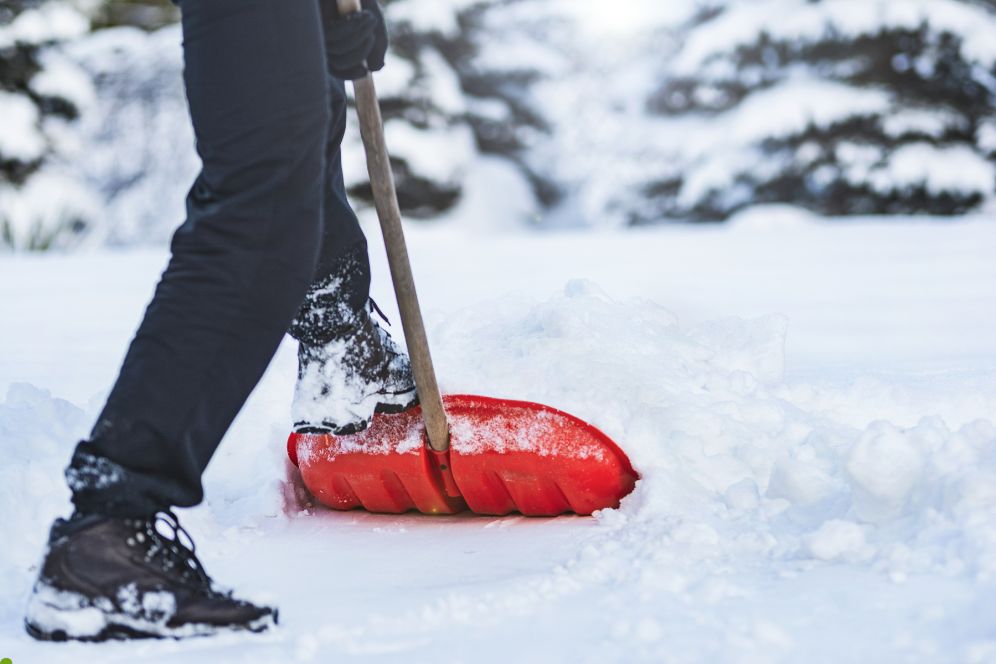 A person shoveling snow with a red shovel, surrounded by a snowy landscape and a clear blue sky.