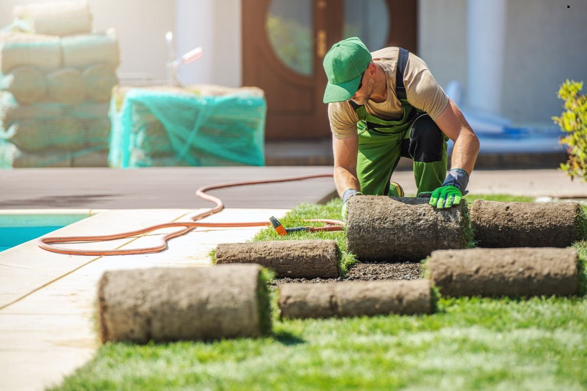 A man in green overalls installs artificial turf while working diligently on a lawn.