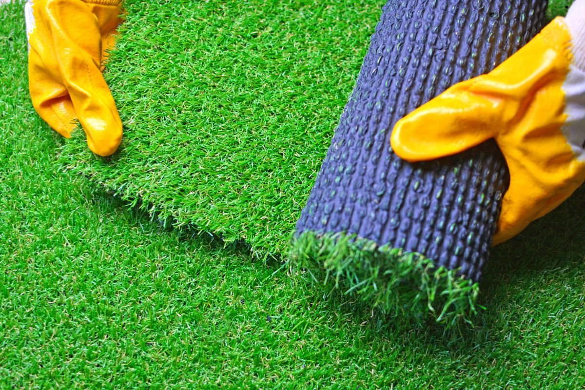 A person with yellow gloves is holding a roll of artificial grass, indicating preparation for an artificial turf installation.